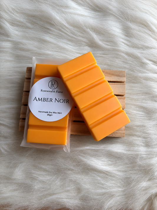 A pack of orange wax melts in a clear plastic bag with a white circular label reading 'Amber Noir' by Rosewood & Flame, placed on a small wooden tray over a white faux-fur surface.