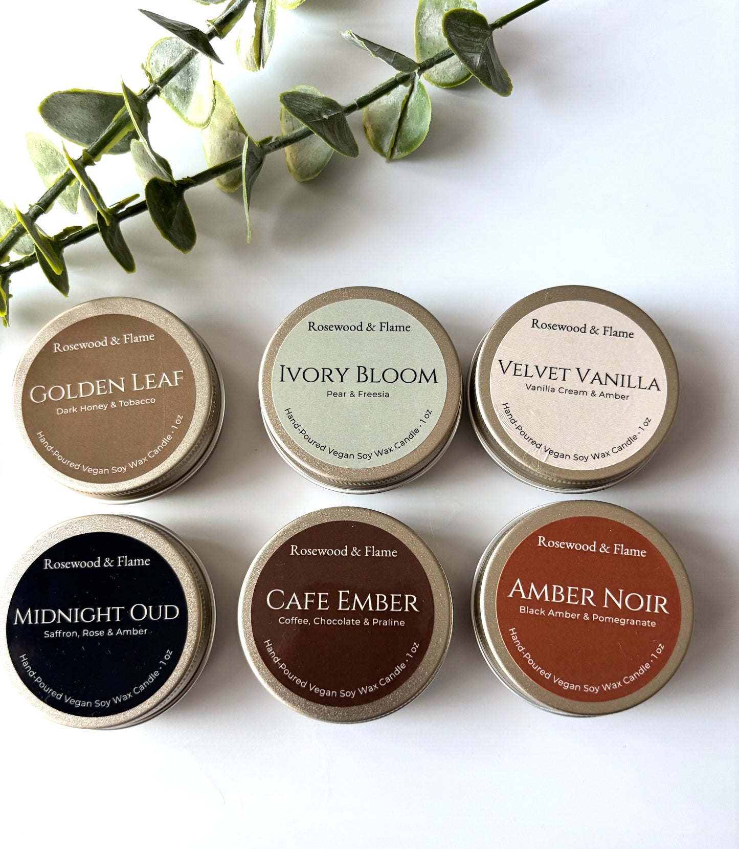 Six round metal soy wax candles in different scents are arranged in two rows on a white background with a green eucalyptus sprig at the top. The scents shown are Golden Leaf, Ivory Bloom, Velvet Vanilla, Midnight Oud, Cafe Ember, and Amber Noir, each with a distinct color label.