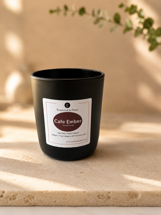 A matte black candle jar labeled 'Cafe Ember' by Rosewood & Flame placed on a light stone surface with warm sunlight and soft shadows.