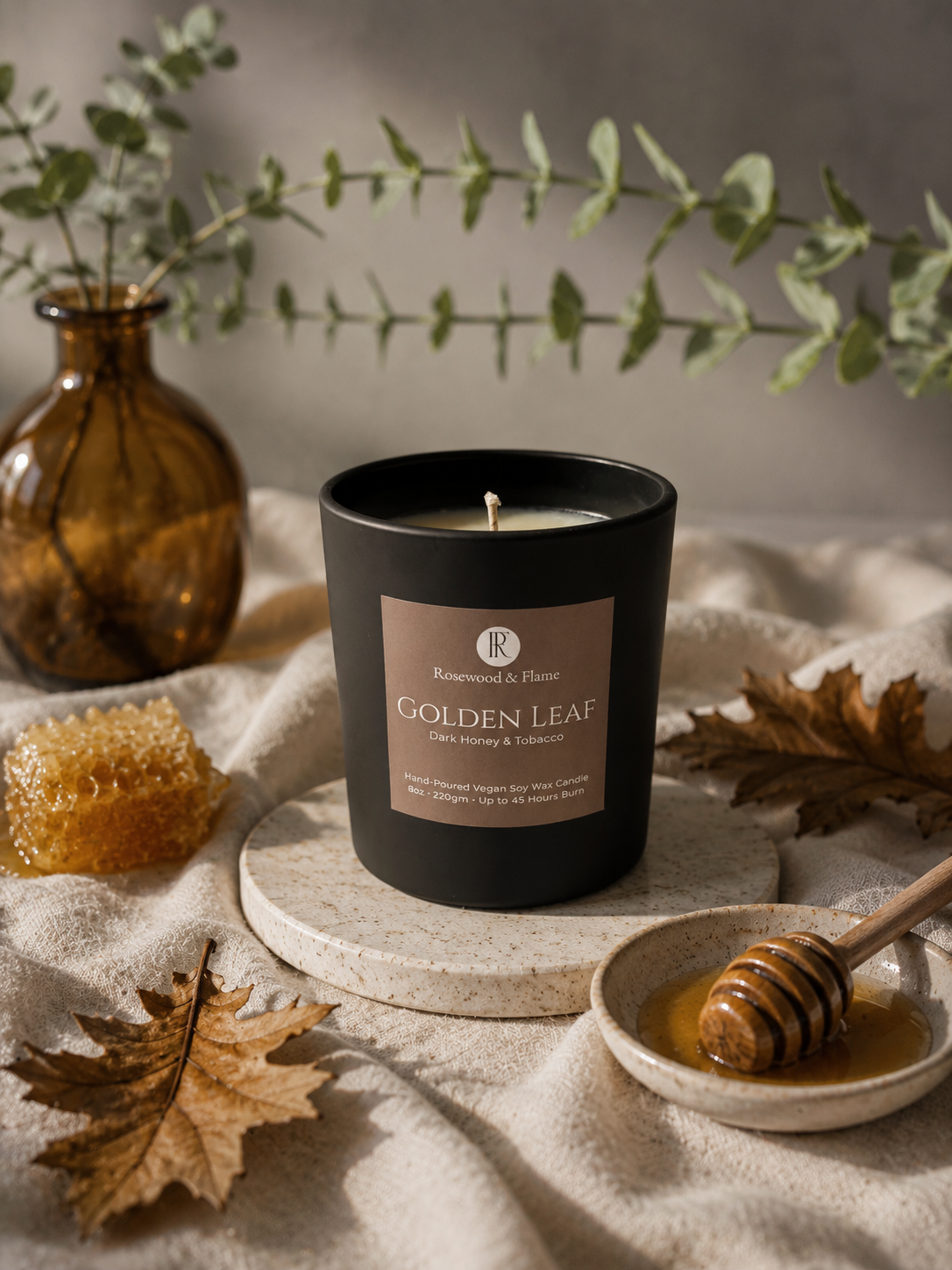 A black candle jar labeled 'Golden Leaf' by Rosewood & Flame, styled on a stone coaster with autumn leaves, honeycomb, a honey dipper in a dish, and a brown glass vase with eucalyptus branches against a neutral background.