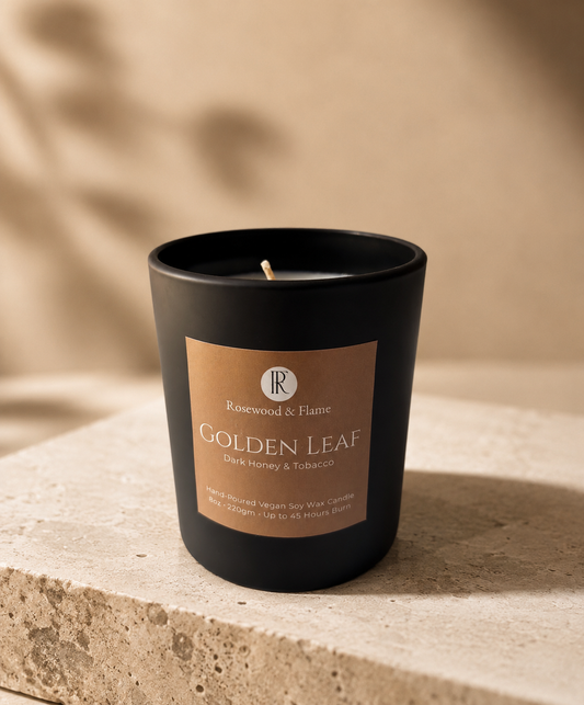 A black glass candle jar with a warm brown label reading 'Rosewood & Flame Golden Leaf Dark Honey & Tobacco,' placed on a textured stone surface against a softly lit beige background.