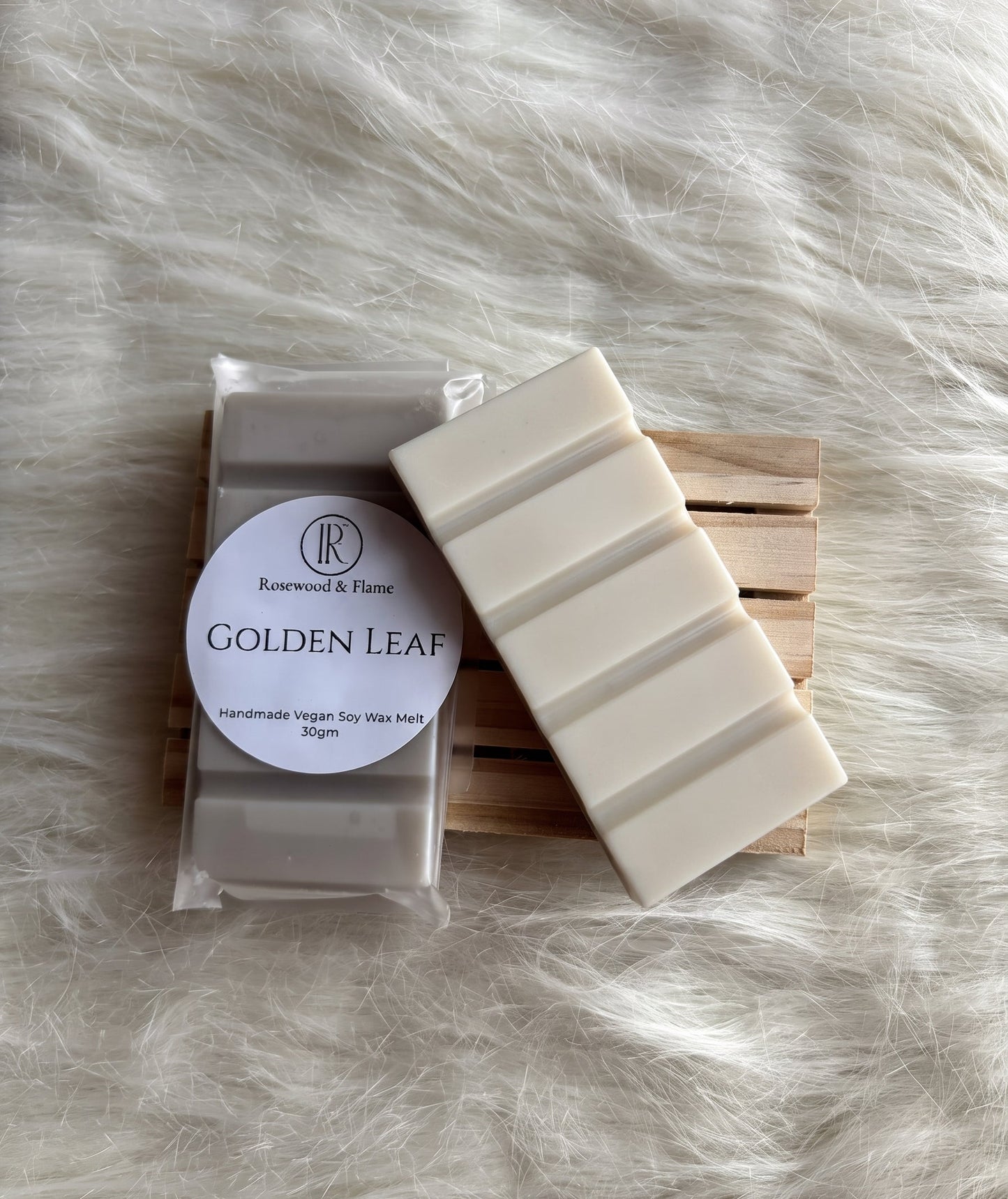 A packaged bar of soap labeled 'Golden Leaf' by Rosewood & Flame, shown next to a stack of uncut cream-colored soap bars on a small wooden stand, placed on a white faux fur surface.