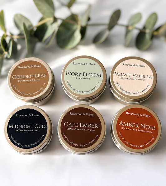 Six small round metal tins of soy wax candles arranged in two rows on a white surface with blurred green leaves in the background. The tins are labeled Golden Leaf, Ivory Bloom, Velvet Vanilla, Midnight Oud, Cafe Ember, and Amber Noir, all under the Rosewood & Flame brand.