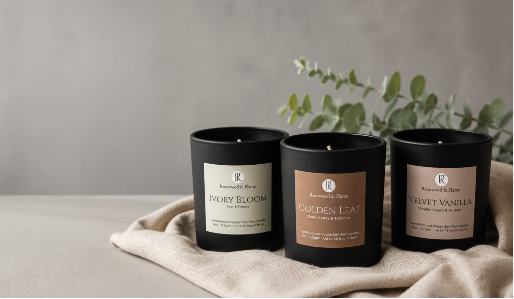 Three black candle jars with cream and tan labels are arranged on a beige cloth against a soft gray wall, with greenery in the background. The labels read Ivory Bloom, Golden Leaf, and Velvet Vanilla.
