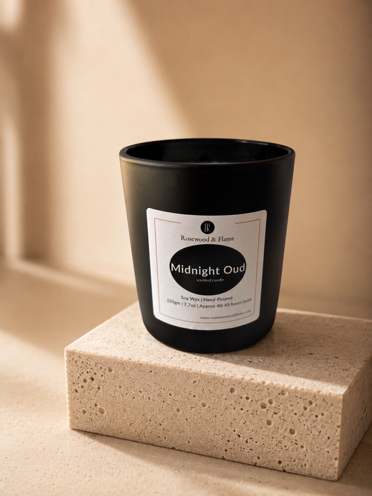 A black glass jar candle labeled 'Midnight Oud' by Rosewood & Flame, displayed on a textured beige stone block against a warm beige background.
