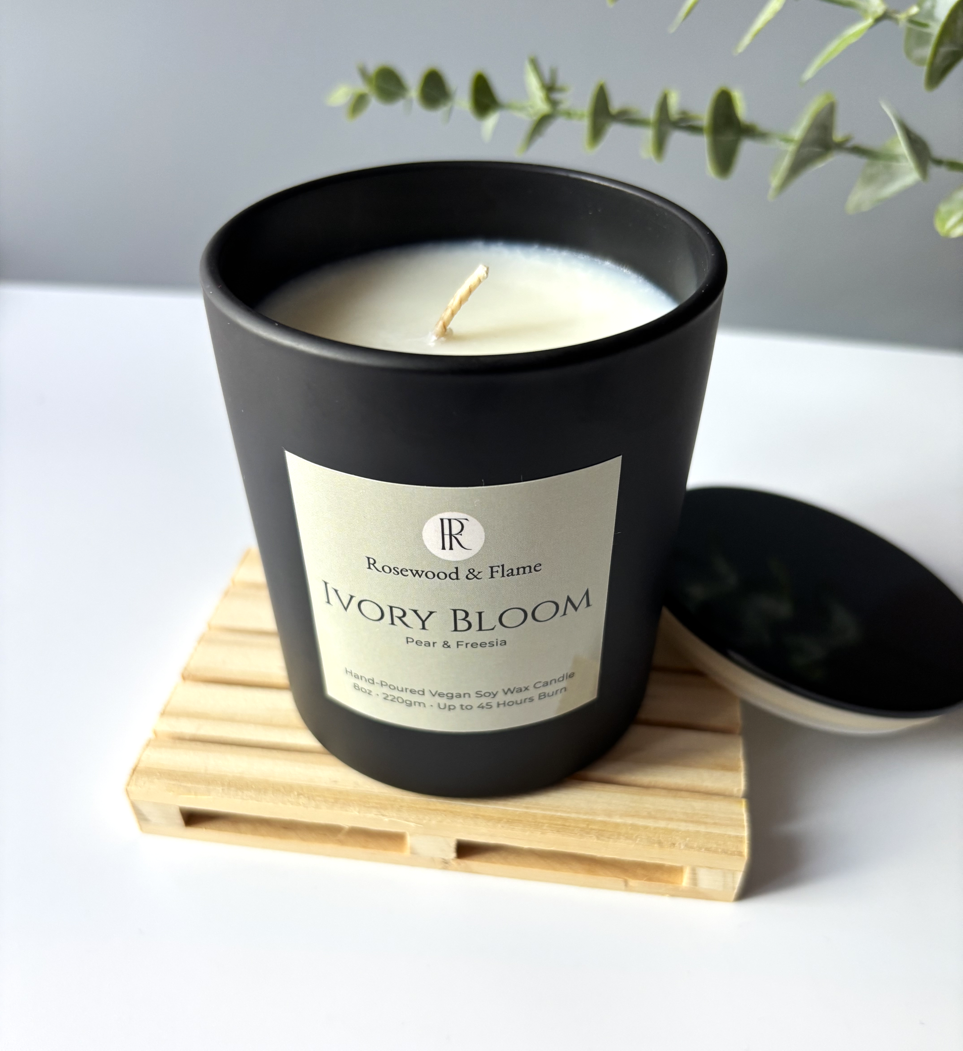 A black ceramic candle jar labeled 'Rosewood & Flame Ivory Bloom' sits on a light wood coaster with its matching lid beside it. The candle has white wax and a single wick, photographed on a white surface with soft greenery in the background.