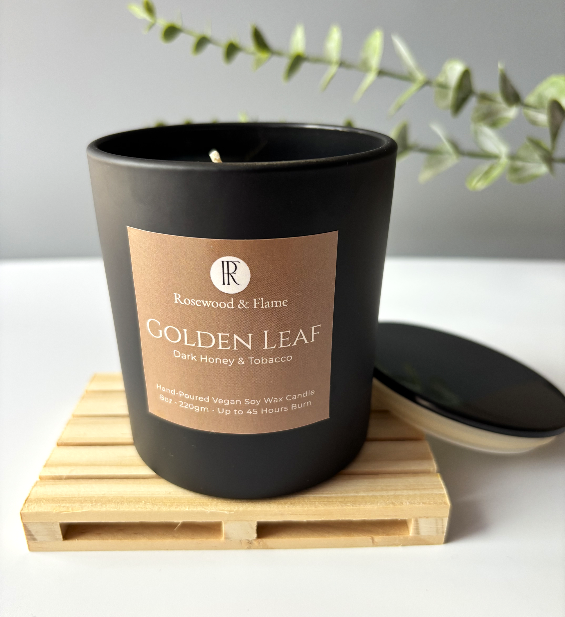 A matte black soy wax candle jar with a rectangular kraft paper label reading 'Rosewood & Flame Golden Leaf Dark Honey & Tobacco,' shown on a light wood tray with its matching lid beside it. A soft-focus green branch appears in the background against a light gray wall.