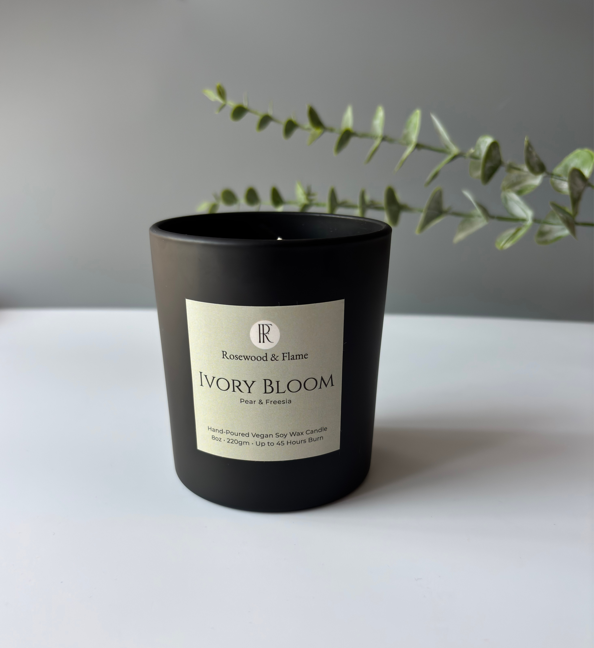 A matte black candle jar with a cream label reading 'Rosewood & Flame' and 'Ivory Bloom', placed on a white surface against a gray wall with a green sprig behind it.