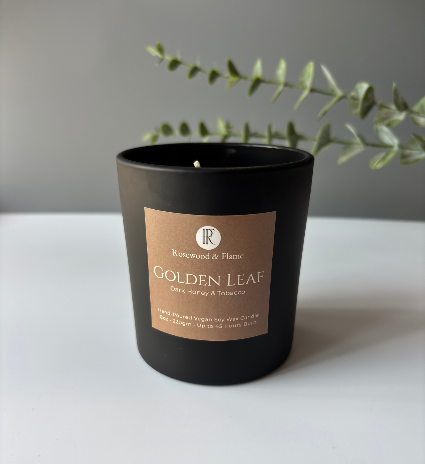 A black glass candle jar with a tan label reading 'Rosewood & Flame Golden Leaf Dark Honey & Tobacco' on a white surface against a gray wall, with green eucalyptus branches behind it.