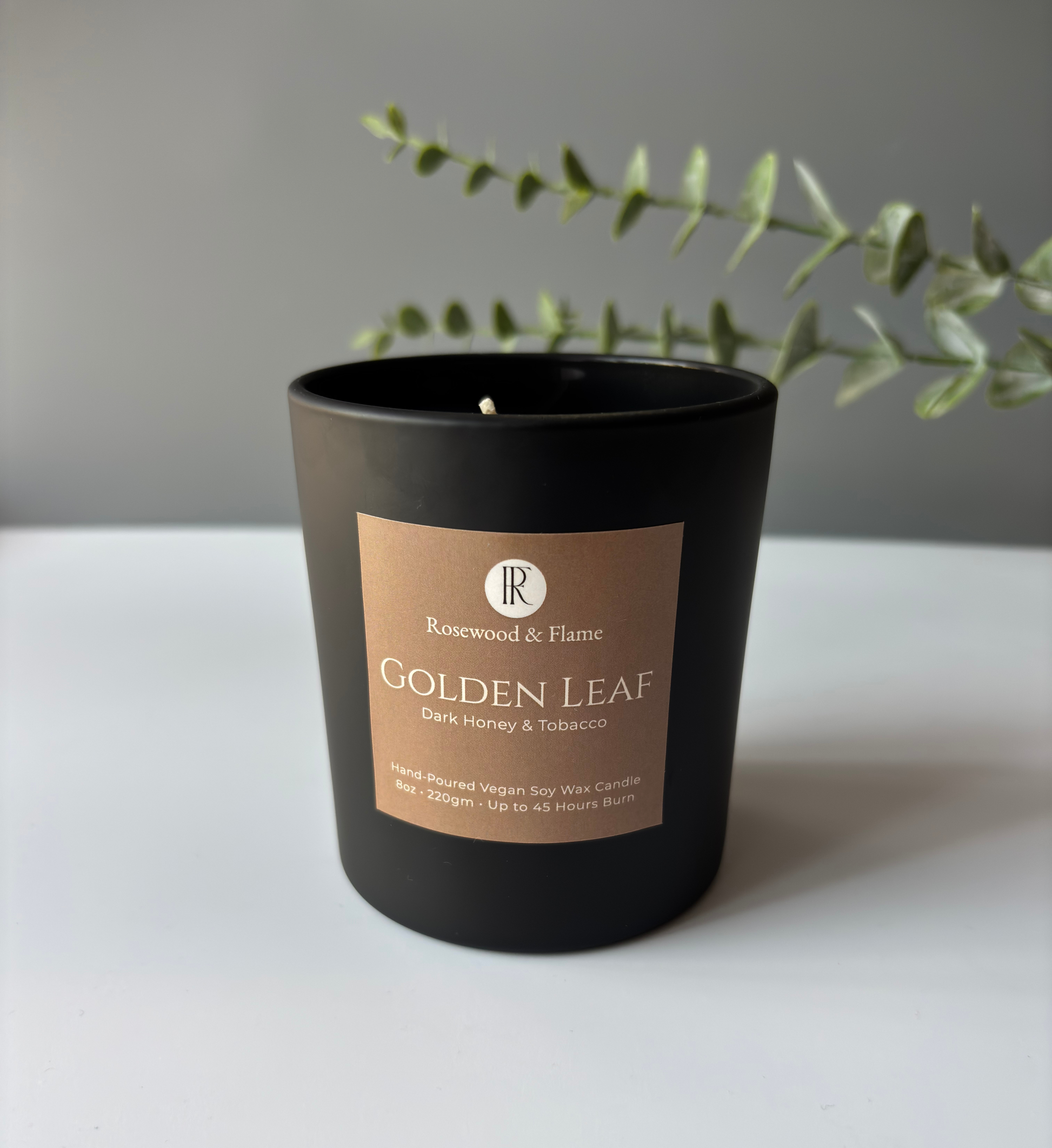A black glass candle jar with a tan label reading 'Rosewood & Flame Golden Leaf Dark Honey & Tobacco' on a white surface against a gray wall, with green eucalyptus branches behind it.
