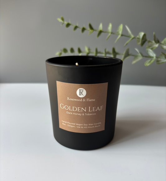 A black glass candle jar with a tan label reading 'Rosewood & Flame Golden Leaf Dark Honey & Tobacco' on a white surface against a gray wall, with green eucalyptus branches behind it.