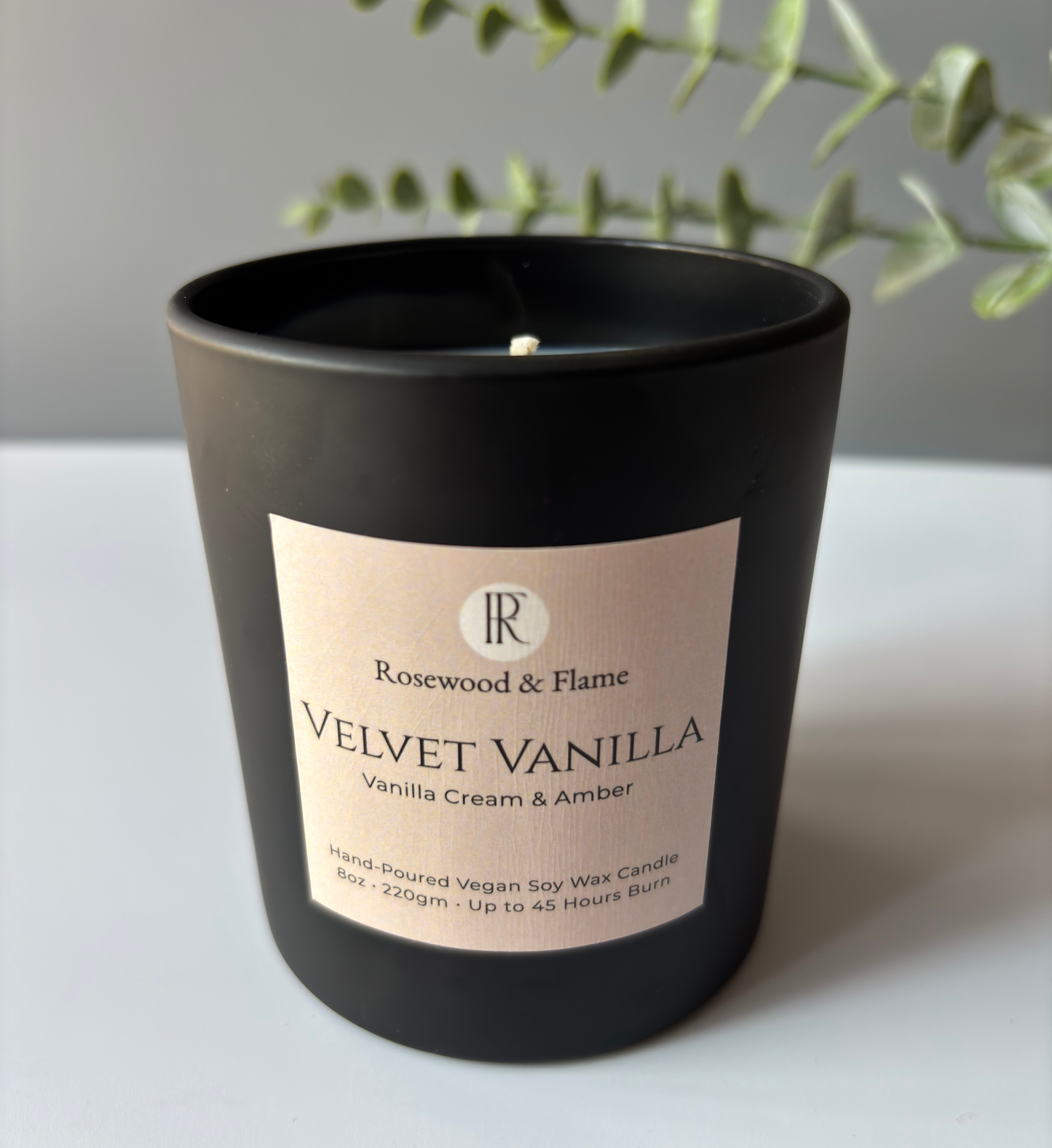A matte black soy wax candle in a cylindrical jar with a cream-colored label reading 'Rosewood & Flame VELVET VANILLA Vanilla Cream & Amber'. The candle is set on a light gray surface with a soft-focus background featuring green foliage.