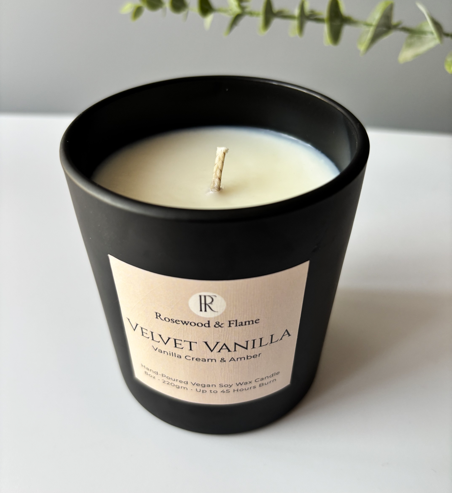 A black candle jar with a cream-colored wax and a beige label reading 'Rosewood & Flame Velvet Vanilla', set on a light surface with a blurred green plant in the background.