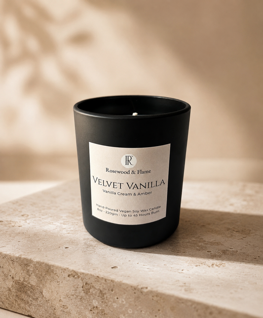 A matte black candle jar with a white label reading 'Rosewood & Flame VELVET VANILLA' and 'Vanilla Cream & Amber', placed on a stone surface against a warm beige background with soft leaf shadows.