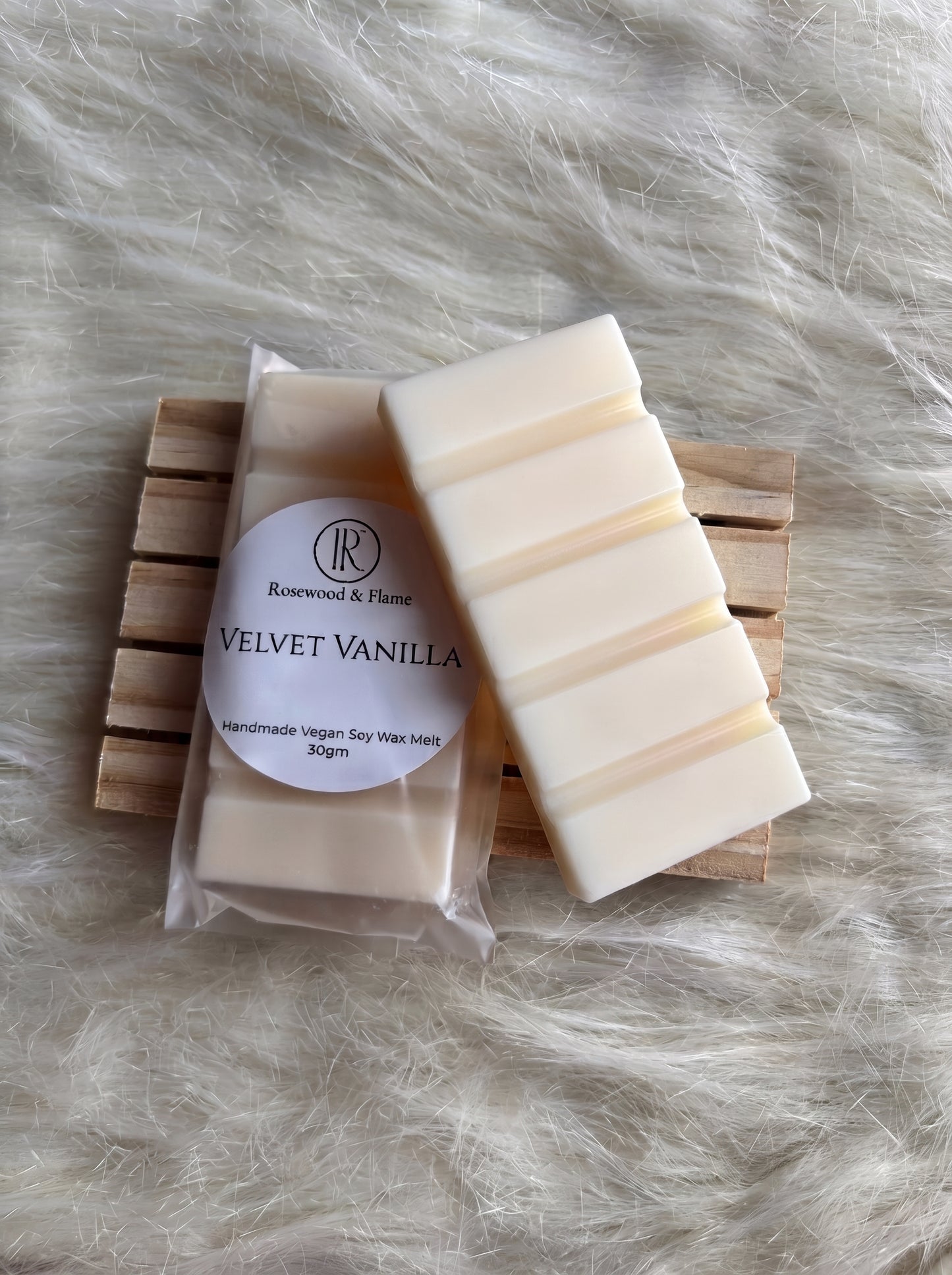 A bar of handmade soy wax melt in its clear plastic packaging with a white label reading 'Rosewood & Flame VELVET VANILLA', placed next to an unwrapped cream-colored wax bar on a small wooden slatted coaster over a white faux fur background.