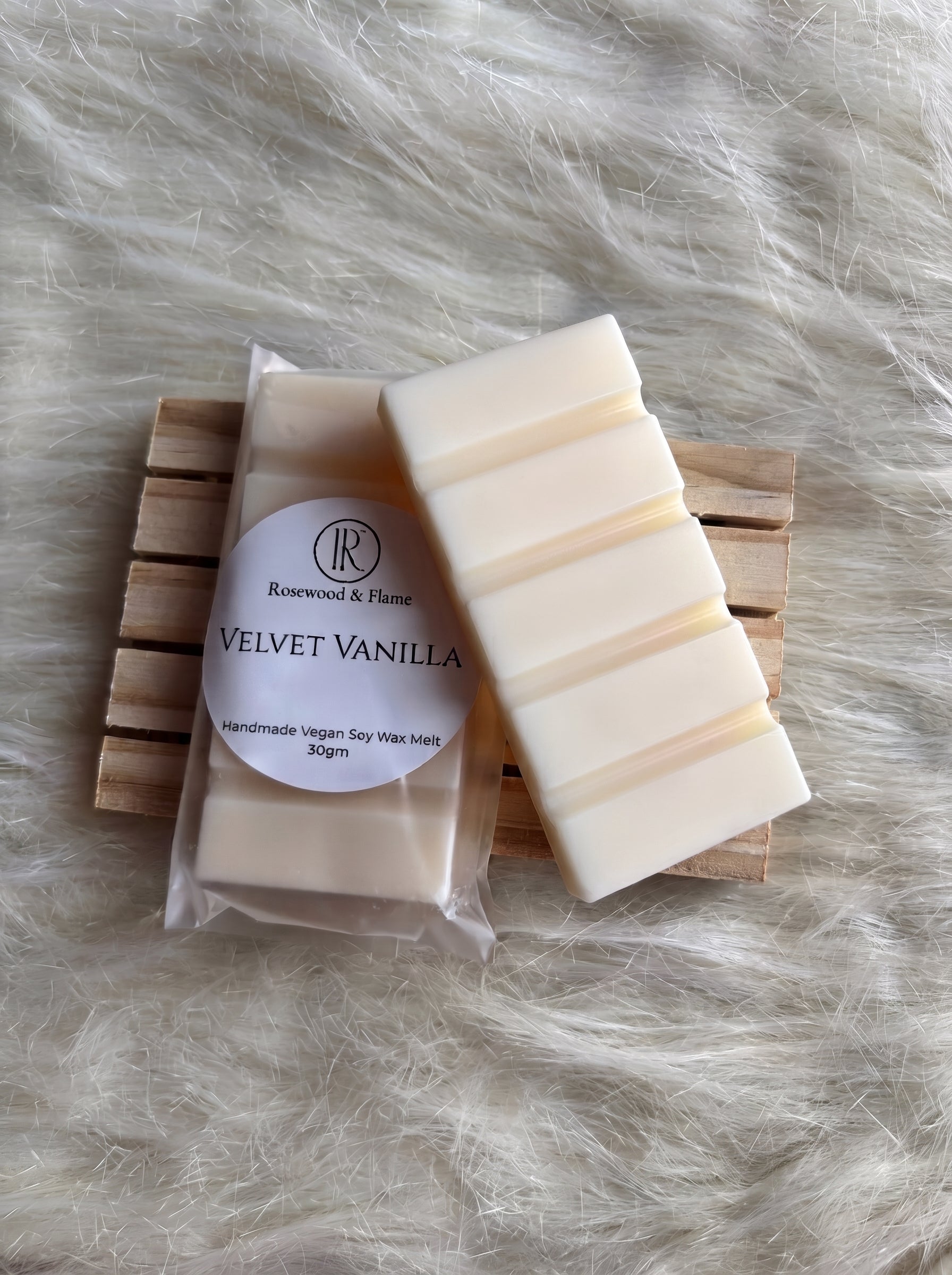 A bar of handmade soy wax melt in its clear plastic packaging with a white label reading 'Rosewood & Flame VELVET VANILLA', placed next to an unwrapped cream-colored wax bar on a small wooden slatted coaster over a white faux fur background.