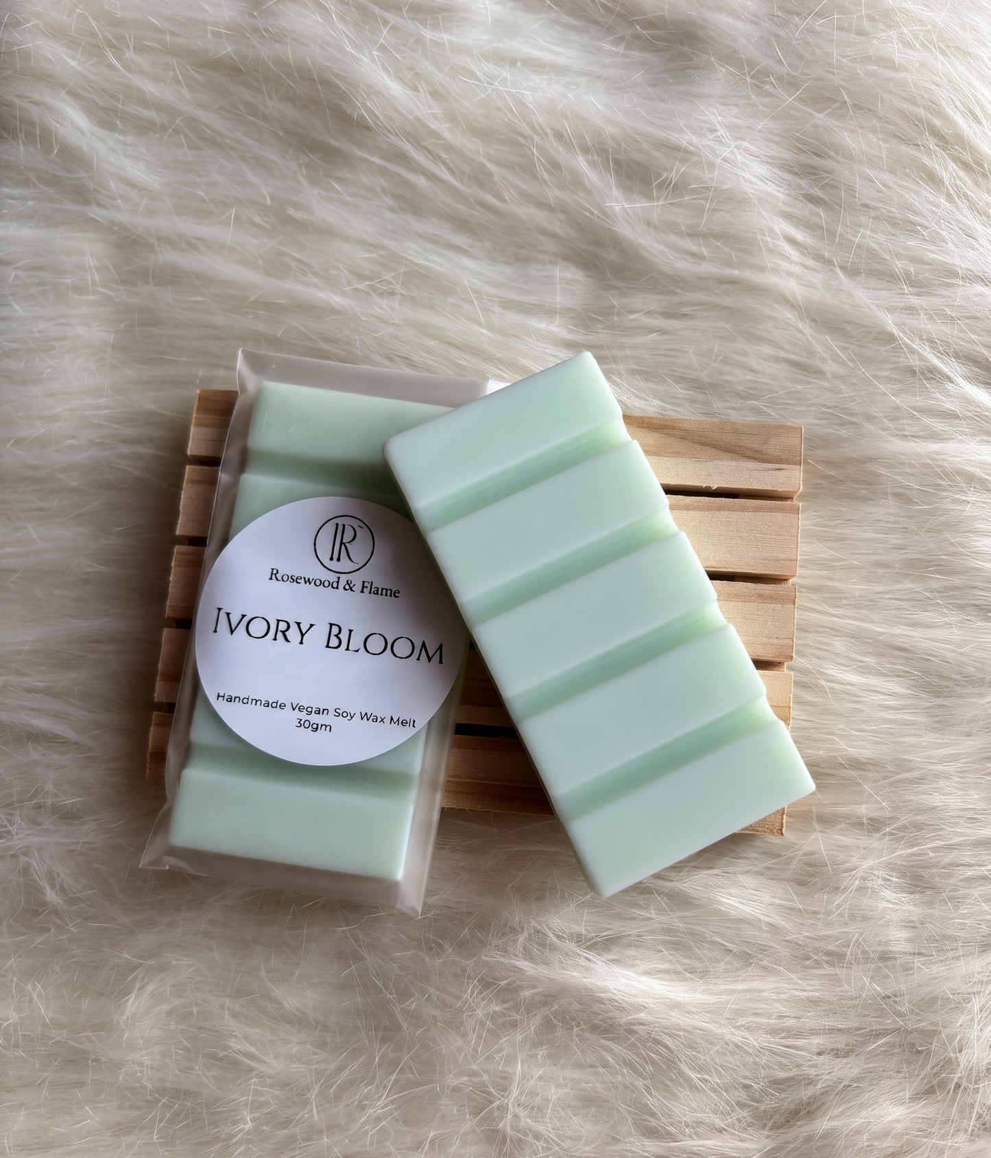 A packaged and unwrapped bar of soap labeled 'Ivory Bloom' by Rosewood & Flame, resting on a wooden slatted coaster over a white faux fur background.