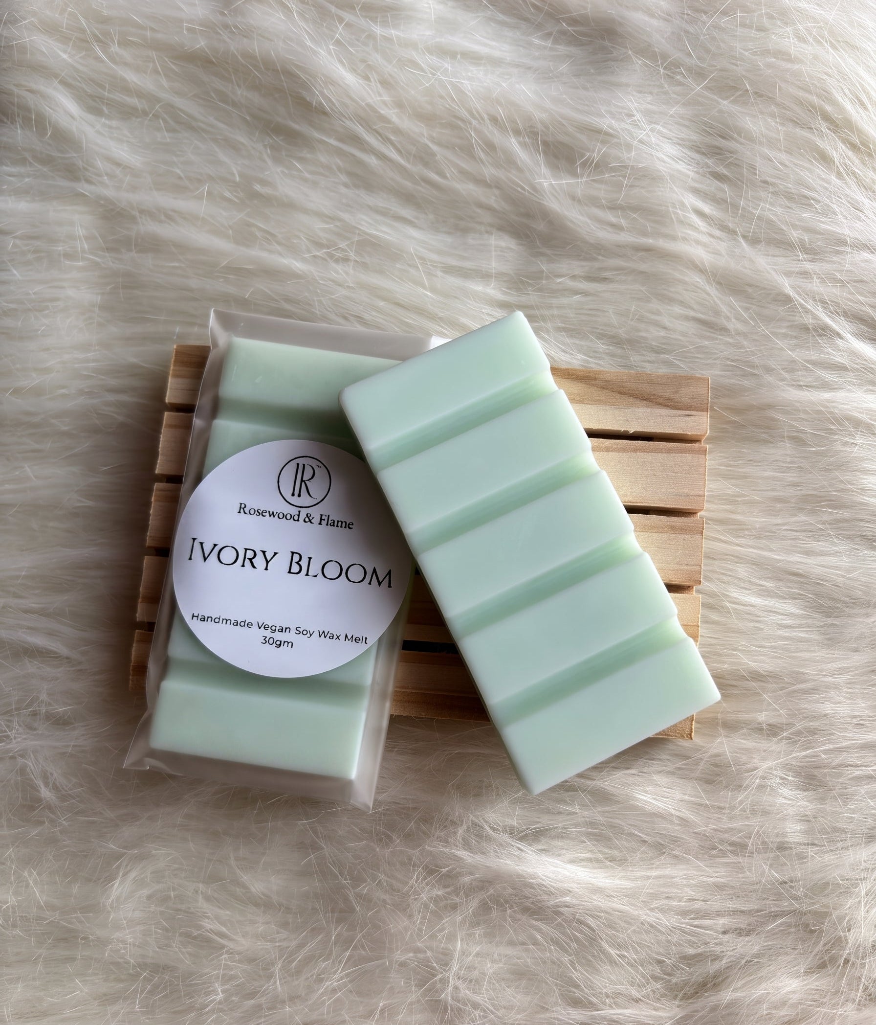 A packaged and unwrapped bar of soap labeled 'Ivory Bloom' by Rosewood & Flame, resting on a wooden slatted coaster over a white faux fur background.