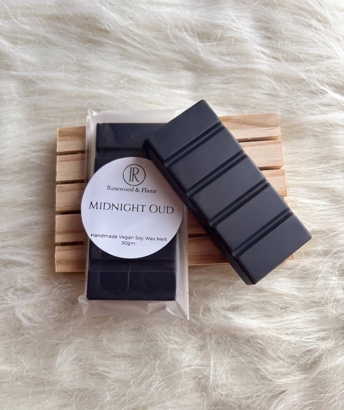 Two dark gray molded wax melt bars on a small wooden slatted tray over a white faux fur background. One bar is in a clear plastic bag with a round white label reading 'Rosewood & Flame' and 'MIDNIGHT OUD'.